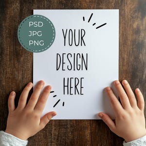 May include: A white sheet of paper with the text "YOUR DESIGN HERE" in black lettering. The paper is held by two hands on a brown wooden surface.