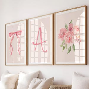 Puede incluir: Tres impresiones artísticas enmarcadas con diseños de acuarela. Las impresiones presentan un lazo rosa, la letra "A" y un ramo floral. La obra de arte está sobre un fondo rosa claro, con marcos de madera.