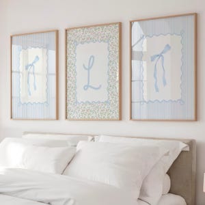 Pode incluir: Três impressões emolduradas com desenhos em azul claro e branco. Duas impressões apresentam um laço azul claro sobre um fundo branco com uma borda listrada azul. A terceira impressão tem uma borda floral e a letra "L" em azul claro.