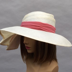 K&ouml;nnte beinhalten: Ein wei&szlig;er Strohhut mit breiter Krempe und einem rosa Band um die Krone. Der Hut hat eine weiche Krempe und ist so konzipiert, dass er vor der Sonne sch&uuml;tzt.