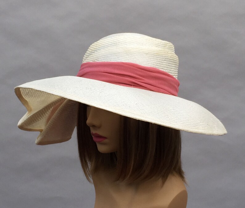 K&ouml;nnte beinhalten: Ein wei&szlig;er Strohhut mit breiter Krempe und einem rosa Band um die Krone. Der Hut hat eine weiche Krempe und ist so konzipiert, dass er vor der Sonne sch&uuml;tzt.