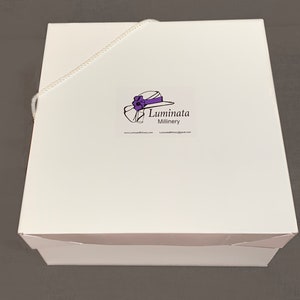 K&ouml;nnte beinhalten: Wei&szlig;e Hutbox mit einem wei&szlig;en Band und einem Logo mit der Aufschrift "Luminata Millinery" mit einer Zeichnung eines Hutes mit einer violetten Blume und einem violetten Band.