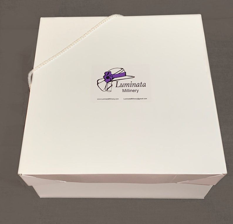 K&ouml;nnte beinhalten: Wei&szlig;e Hutbox mit einem wei&szlig;en Band und einem Logo mit der Aufschrift "Luminata Millinery" mit einer Zeichnung eines Hutes mit einer violetten Blume und einem violetten Band.