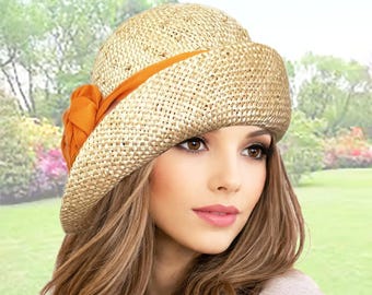 May include: A straw hat with a rounded crown and a wide, downward-sloping brim. The hat features an orange ribbon and a decorative flower on the side. The hat is a light tan color and appears to be made of woven straw.