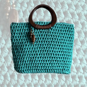 May include: A turquoise crochet handbag with a dark brown wooden handle. The bag features a rectangular shape and a decorative wooden bead charm. The crochet pattern is visible, and the bag appears to be handmade.