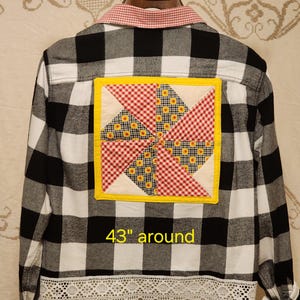 May include: A black and white buffalo plaid button-down shirt featuring a red and white gingham collar, a white lace hem, and a square quilt patch on the back with a yellow border and a pinwheel pattern. The shirt measures 43 inches around.