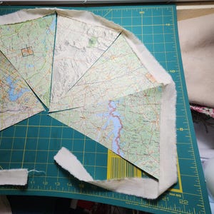 May include: A partially constructed craft project featuring map segments with a cream-colored fabric border. The maps display green and blue geographical features. The project is on a green cutting mat with measurement markings.