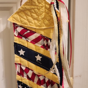 Puede incluir: Decoración de tela hecha a mano con un techo acolchado amarillo y un cuerpo con rayas rojas y blancas, secciones azules con estrellas blancas y paneles amarillos. Cintas doradas, rojas y azules cuelgan del costado. Suspendido por una cuerda marrón.