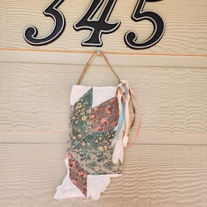以下が含まれることがあります： インディアナ州の形をしたキルトの壁掛け。緑、茶色、白の色合いの花柄と幾何学模様が特徴です。ジュートロープで吊り下げられ、カラフルなリボンで飾られています。上部には、家の番号「345」が表示されています。