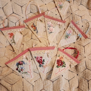 Könnte beinhalten: Eine Sammlung dreieckiger Stoffbanner mit Blumenmustern und dem Wort "Liebe". Jedes Banner hat eine rosa Umrandung, Spitzendetails und einen Griff. Die Banner sind auf einer cremefarbenen, gehäkelten Decke angeordnet.