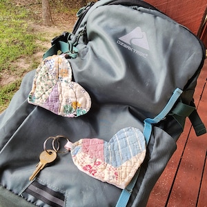 Puede incluir: Una mochila gris Ozark Trail con dos corazones de tela acolchada. Un corazón está sujeto al asa superior, mientras que el otro está conectado a un llavero de metal con una llave de latón. Los corazones presentan patrones de patchwork en tonos pastel.