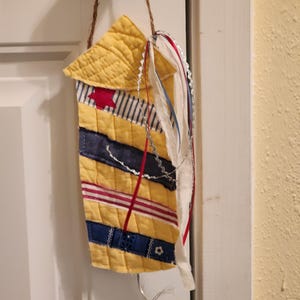 May include: A handcrafted patriotic wall decoration. The quilted fabric features yellow, blue, and red stripes and a red star. Decorative ribbons and a twine loop for hanging complete the design. The hanging is attached to a white door.