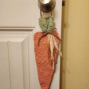 Puede incluir: Un colgador de puerta con forma de zanahoria acolchada, de color melocotón, con una parte superior floral verde y un lazo de yute. La zanahoria tiene un patrón de diamantes. Plumas y rafia están unidas al lazo.