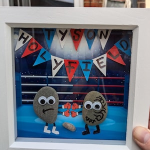 Può includere: Un'opera d'arte incorniciata con due figure di pietra dipinte in un ring di boxe. Le pietre hanno occhi sporgenti e indossano guantoni da boxe. L'opera ha uno sfondo blu con una ghirlanda rossa e blu che recita "TYSON HOLYFIELD".