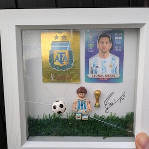 Może przedstawiać: Biała rama z pękniętym szklanym panelem. Wewnątrz ramy znajduje się figurka Lego Lionela Messiego w niebiesko-białej koszulce piłkarskiej, piłka nożna, złoty puchar i dwie karty kolekcjonerskie. Karty kolekcjonerskie przedstawiają Lionela Messiego i herb argentyńskiej reprezentacji narodowej. Rama zawiera również podpis, który wydaje się być "Messi".