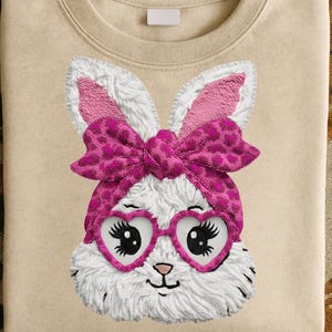 Può includere: Felpa beige con un disegno a forma di coniglio bianco. Il coniglio ha orecchie rosa, un fiocco e occhiali a forma di cuore. Il disegno è al centro della felpa, con una consistenza soffice e stampa leopardata rosa.