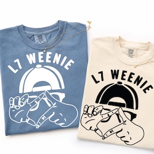 May include: Two t-shirts, one blue and one cream-colored, with the text "L7 Weenie" in white and black, respectively. The design includes a baseball cap and hand gestures. Both shirts have a Comfort Colors tag.