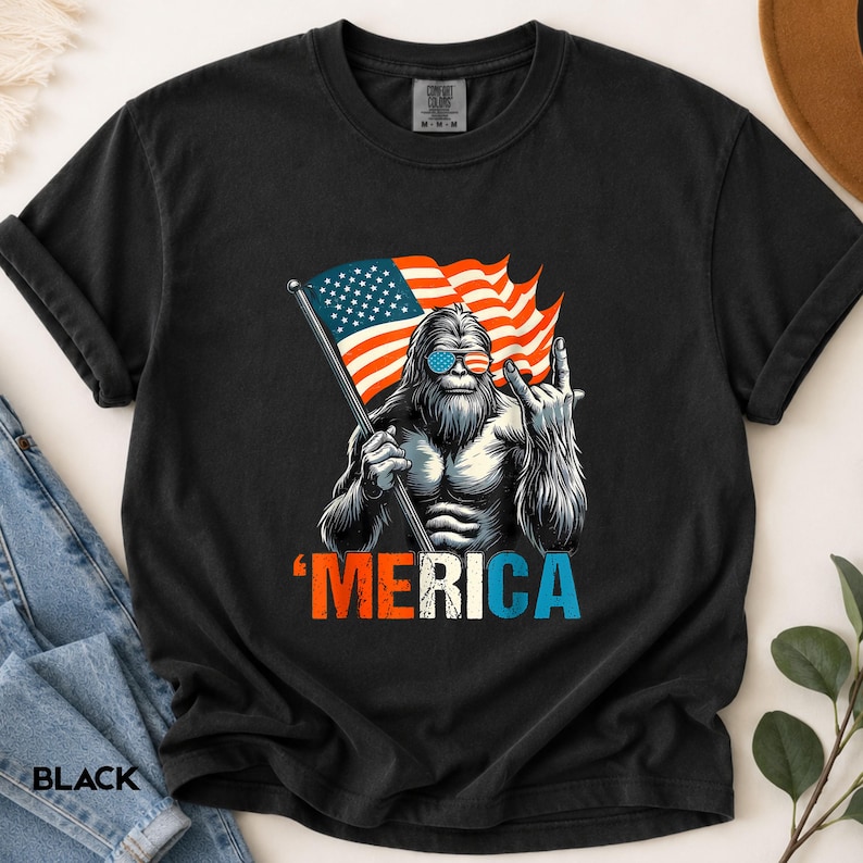 Puede incluir: Camiseta negra con un gr&aacute;fico de una criatura parecida a Bigfoot con gafas de sol y sosteniendo una bandera estadounidense. La palabra "'MERICA" est&aacute; impresa en rojo, blanco y azul. La camiseta est&aacute; hecha de un material suave.