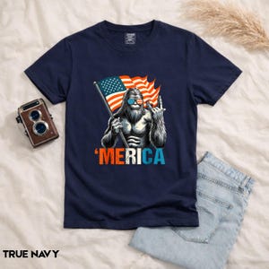 Puede incluir: Camiseta azul marino con un gr&aacute;fico de una criatura parecida a Pie Grande con gafas de sol y una bandera estadounidense. La palabra "'MERICA" est&aacute; impresa en rojo y azul. La camiseta se combina con vaqueros azules.