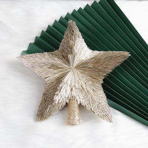 May include: A gold star-shaped Christmas tree topper with a glittery gold base. The star is made of many small, gold-colored pieces radiating from the center. A dark green, folded paper fan is in the background.