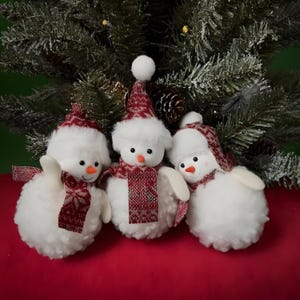 Puede incluir: Tres muñecos de nieve de peluche con cuerpos blancos, narices naranjas y ojos de botón negros. Cada muñeco de nieve lleva un gorro y una bufanda estampados en rojo y blanco. Los muñecos de nieve están colocados frente a un árbol de Navidad.