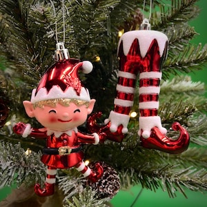May include: Two Christmas tree ornaments: a smiling elf figure and a pair of elf legs. The elf is red with white trim, a black belt, and a gold buckle. The legs are red and white striped with white trim.