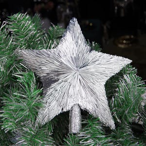 May include: A silver star tree topper with a glittery base, set against a backdrop of green and silver Christmas tree branches. The star is made of many silver rods radiating from the center.