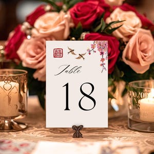 May include: A table number card with the number 18 in black, featuring floral and bird illustrations. The card is set on a table with candles and a floral arrangement of red and pink roses.
