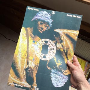 Peut inclure: Une affiche rectangulaire présentant le portrait d'une personne portant un bonnet bleu texturé et des lunettes, vêtue d'un haut jaune à motifs. Le texte Alpha Wann et Paris 14e Arr. figure en haut et en bas, avec un logo circulaire central DON DA DADA.