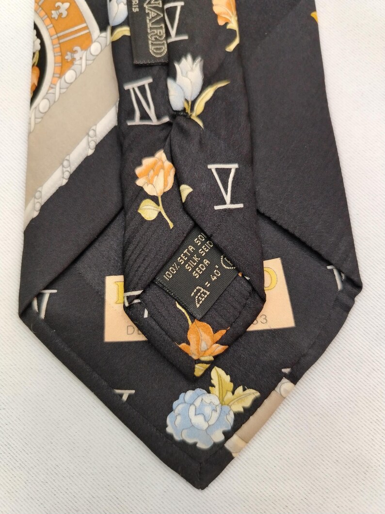 May include: Black silk tie with floral and Roman numeral designs. The tie features orange, white, and blue flowers, along with the Roman numerals IV and V. The label reads "100% Seta 501 Silk Seid = 40'".