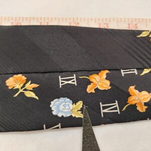 May include: Black tie with a floral pattern in orange and blue. The tie has a striped texture and Roman numerals. A measuring tape is placed over the tie.
