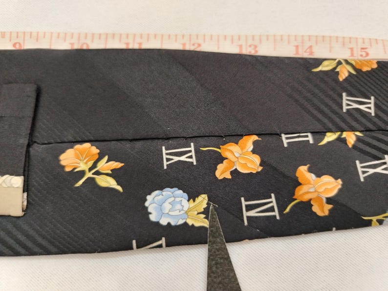 May include: Black tie with a floral pattern in orange and blue. The tie has a striped texture and Roman numerals. A measuring tape is placed over the tie.