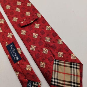 May include: Red silk tie with a repeating square pattern and the Burberrys of London label. The tie features a classic plaid design at the tip and a small square pattern throughout the body.