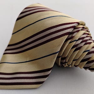 May include: A rolled-up necktie with a cream-colored background and burgundy, navy, and white stripes. The tie is made of a smooth, possibly silk, material. The stripes run horizontally across the tie.