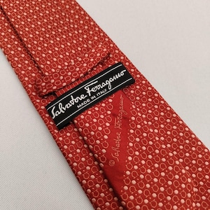 May include: A red silk tie with a repeating pattern of small white circles. The tie features a black label with the text "Salvatore Ferragamo MADE IN ITALY". The brand name is also printed on the tie.
