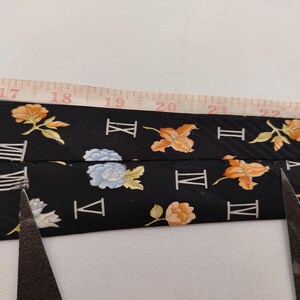 May include: A black tie with a floral and Roman numeral pattern is measured with a tape measure. The tie features orange, yellow, and blue flowers. Scissors are visible, suggesting the tie is being cut.