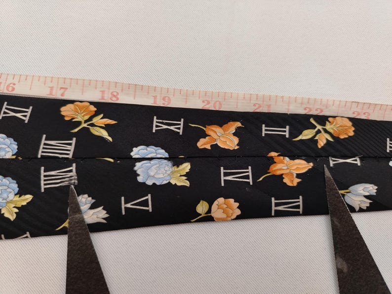 May include: A black tie with a floral and Roman numeral pattern is measured with a tape measure. The tie features orange, yellow, and blue flowers. Scissors are visible, suggesting the tie is being cut.