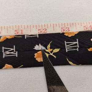 May include: Black necktie with a floral and Roman numeral pattern. The tie is being measured with a tape measure. The tie has orange and white floral designs and white Roman numerals.