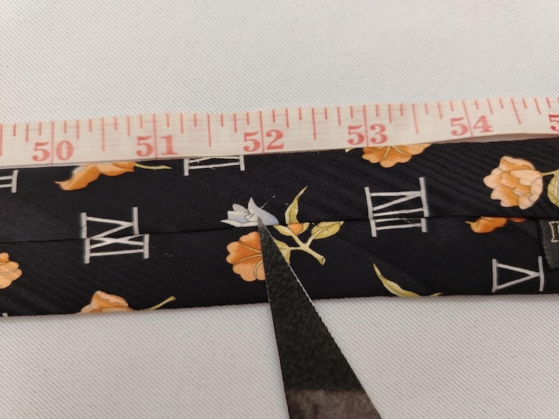 May include: Black necktie with a floral and Roman numeral pattern. The tie is being measured with a tape measure. The tie has orange and white floral designs and white Roman numerals.