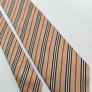 May include: A tan necktie with a striped pattern. The stripes include black, white, and red vertical lines. The tie is made of a smooth fabric and is displayed on a white surface.