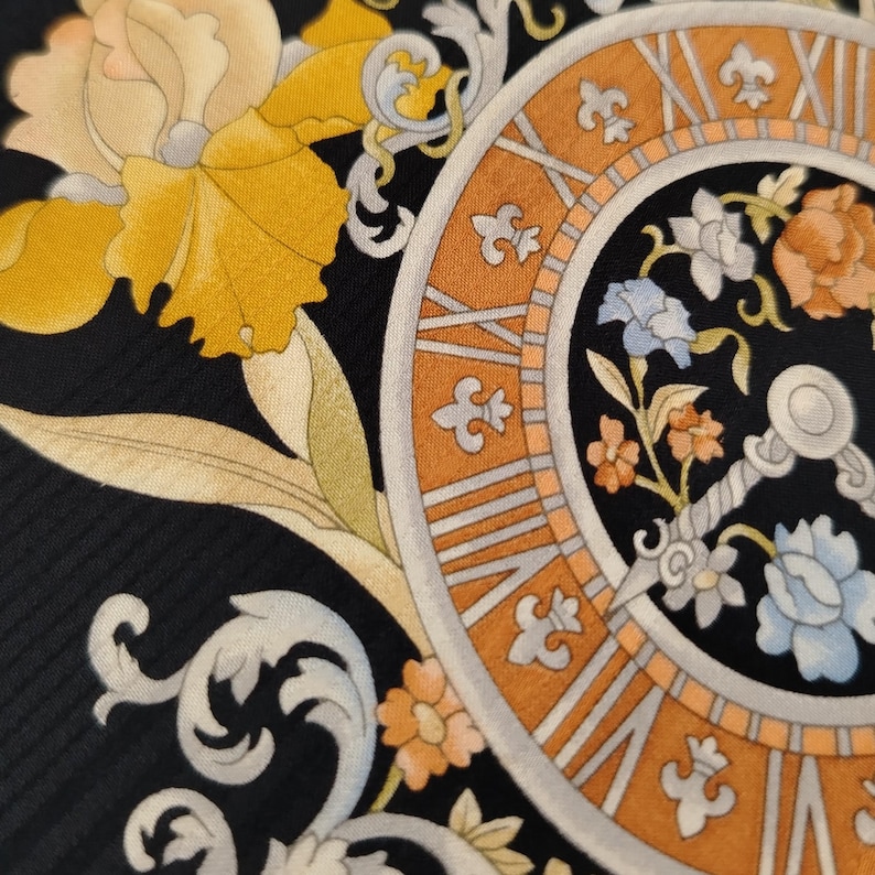 May include: Close-up of a black silk scarf with a clock design. The clock face is orange and silver with Roman numerals. Floral and leaf patterns in yellow, orange, and white surround the clock.