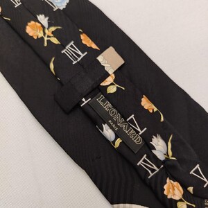 May include: Black silk tie with a floral and Roman numeral pattern. The tie features a repeating design of orange, white, and blue flowers, along with white Roman numerals. The label reads "LEONARD PARIS".
