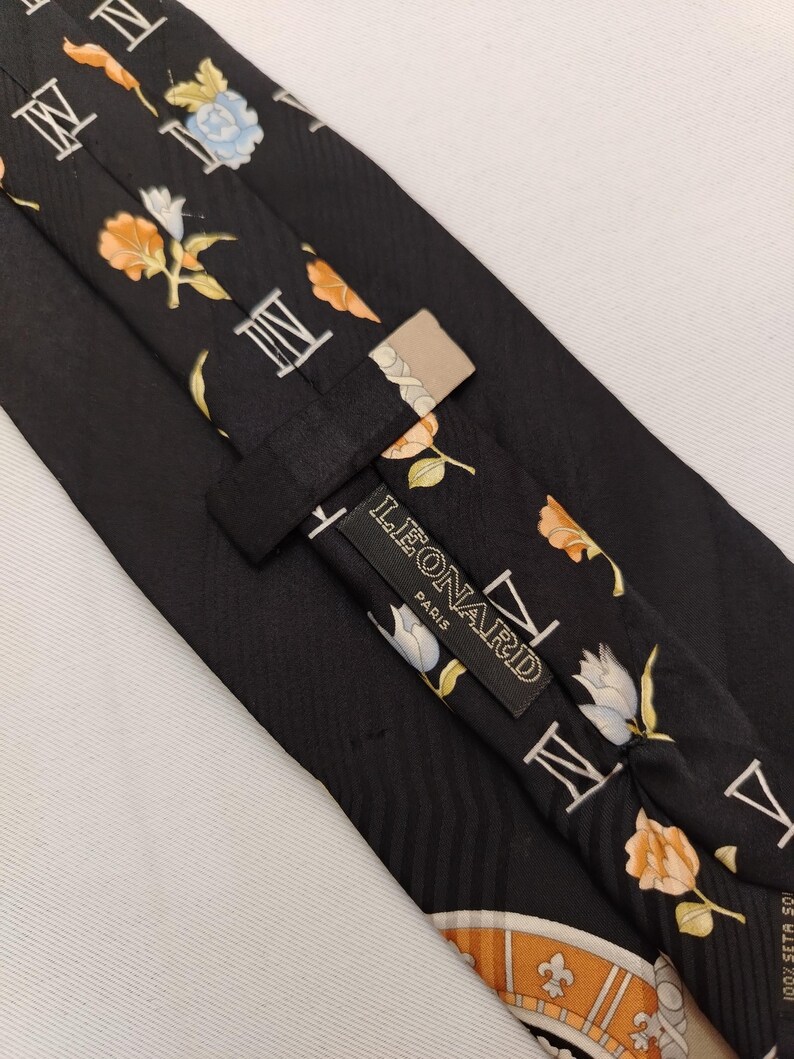 May include: Black silk tie with a floral and Roman numeral pattern. The tie features a repeating design of orange, white, and blue flowers, along with white Roman numerals. The label reads "LEONARD PARIS".