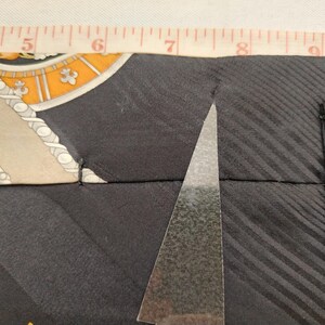 May include: Close-up of a black silk tie with a patterned design in beige, orange, and white. A silver triangle-shaped object is positioned on the tie. A measuring tape shows the tie's width. The tie's design includes diagonal stripes.