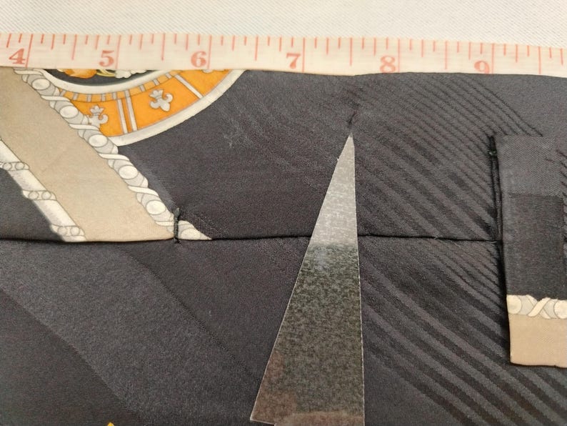 May include: Close-up of a black silk tie with a patterned design in beige, orange, and white. A silver triangle-shaped object is positioned on the tie. A measuring tape shows the tie's width. The tie's design includes diagonal stripes.