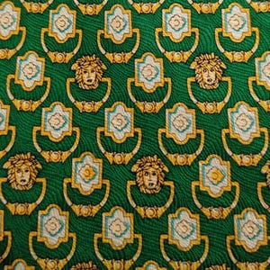 Puede incluir: Tela verde con un patrón repetido de cabezas de Medusa doradas y diseños de aldabas ornamentadas. Las aldabas tienen un diseño floral blanco y azul claro en el centro.