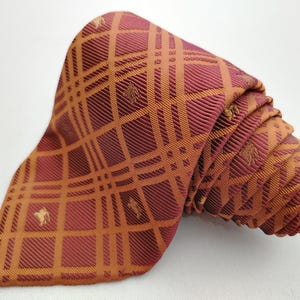 May include: Rolled-up tie with a plaid pattern in shades of burgundy and orange. The tie features a repeating pattern of small horse silhouettes within the plaid design. The tie is made of a shiny, possibly silk, material.