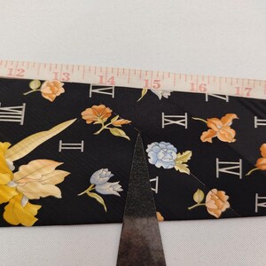 May include: Black fabric with a floral and Roman numeral pattern. The flowers are in shades of yellow, orange, and blue. A ruler is placed over the fabric, showing measurements from 12 to 18 inches.