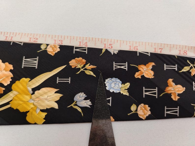 May include: Black fabric with a floral and Roman numeral pattern. The flowers are in shades of yellow, orange, and blue. A ruler is placed over the fabric, showing measurements from 12 to 18 inches.