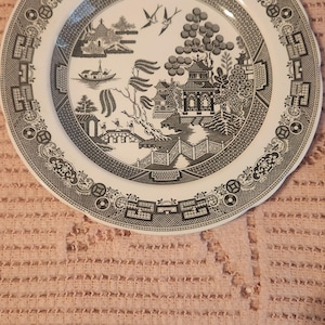May include: A black and white willow pattern dinner plate with a decorative border. The plate features a traditional Chinese landscape scene with a pagoda, willow tree, and birds.